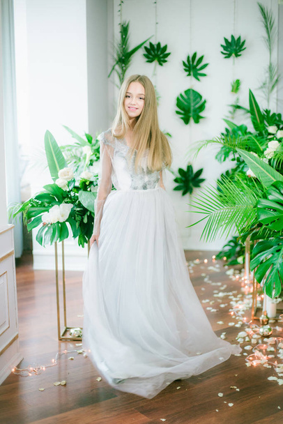 Attractive young girl with blond hair and evening make-up in a romantic dress among tropical greenery and white orchids, roses and lilies. Beauty and fashion - Foto, afbeelding
