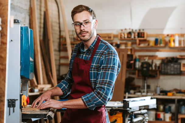 Selektiver Fokus eines gutaussehenden Mannes mit Brille und Schürze in der Werkstatt  - Foto, Bild