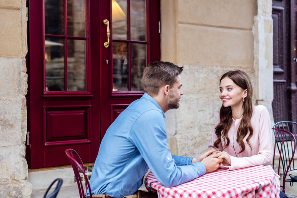 happy young couple holding hands and looking at each other while sitting in street cafe - Photo, Image