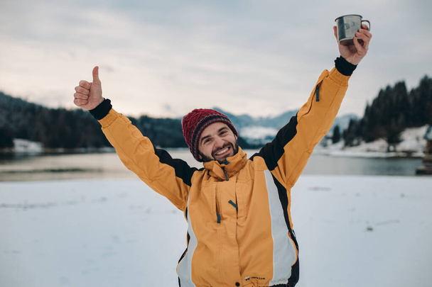Portrait of smiling large man in front of the camera , in the middle of landscape with amazing background of lake and snowy forest and mountain, showing a big like - Photo, Image