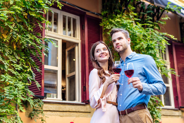 happy couple holding glasses of red wine while standing near house covered with green ivy and looking away - Fotó, kép