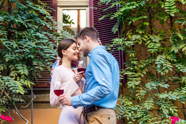 happy couple embracing with closed eyes and holding glasses of red wine while standing near house covered with green ivy - Foto, afbeelding