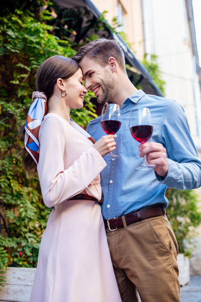 happy couple embracing while standing face to face and holding glasses of red wine - Photo, Image