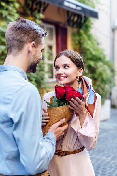 young man presenting bouquet of roses to happy girlfriend on street - Φωτογραφία, εικόνα
