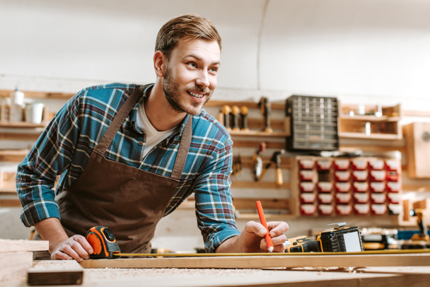 selective focus of cheerful bearded woodworker holding pencil near measuring tape in workshop  - 写真・画像