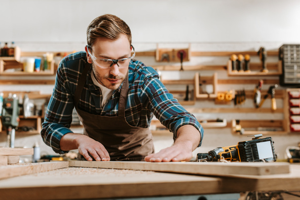 επιλεκτική εστίαση του όμορφου ξυλουργού σε γυαλιά αγγίζοντας ξύλινη σανίδα  - Φωτογραφία, εικόνα