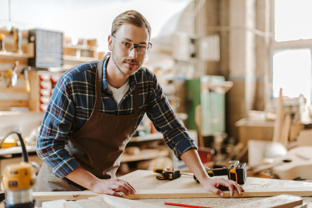 selective focus of handsome carpenter in goggles standing near wooden plank on table  - Photo, Image