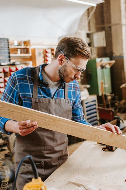 selective focus of handsome carpenter in goggles holding wooden plank in workshop  - Foto, Imagem