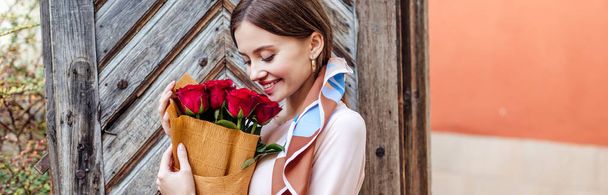 happy girl holding bouquet of red roses while standing near old wooden door - Foto, afbeelding