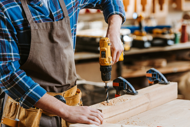 vista recortada del carpintero en delantal sosteniendo taladro de martillo cerca de tablones de madera
 - Foto, Imagen