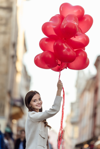 happy girl looking at camera while holding bunch of red heart-shaped balloons on street - Photo, Image