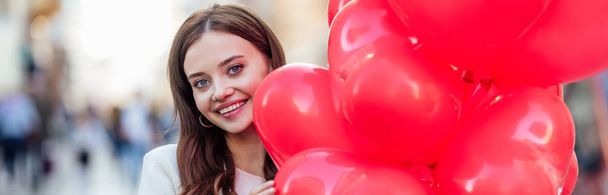 panoráma lövés gyönyörű lány mosolyog a kamera, miközben kezében köteg piros szív alakú lufik - Fotó, kép