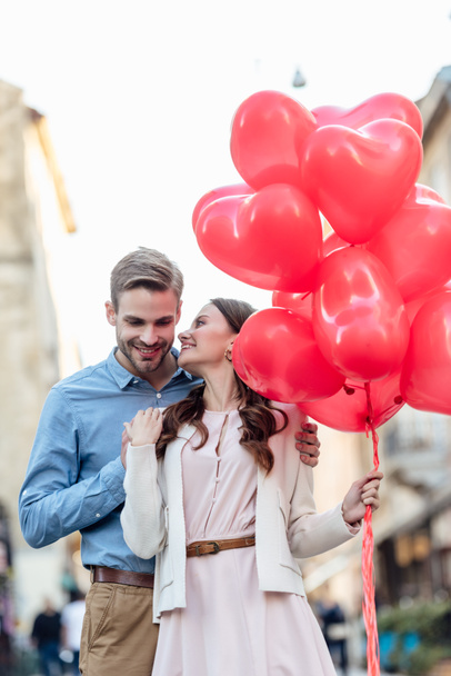 uśmiechnięty mężczyzna obejmujący szczęśliwą dziewczynę trzymając czerwone balony w kształcie serca na ulicy - Zdjęcie, obraz