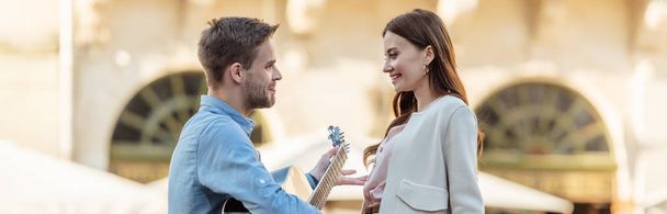 πανοραμική λήψη του όμορφου άντρα που παίζει κιθάρα για την όμορφη κοπέλα του στο δρόμο - Φωτογραφία, εικόνα