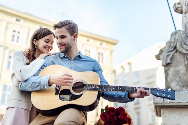 happy girl leaning on shoulder of boyfriend playing acoustic guitar on street - Фото, изображение