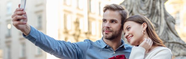 panoramic shot of handsome man taking selfie on smartphone with happy girlfriend - Photo, Image