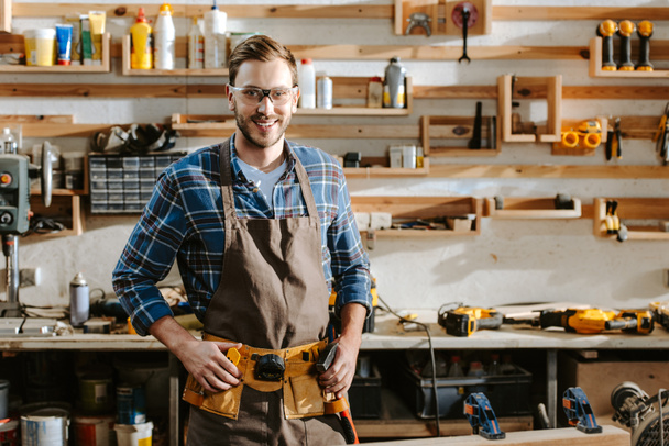 carpintero alegre en gafas de pie y tocando el cinturón de herramientas en el taller
  - Foto, Imagen