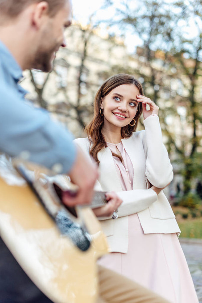 selektywne skupienie się na ładnej, szczęśliwej dziewczynie patrzącej na chłopaka grającego na gitarze na ulicy - Zdjęcie, obraz