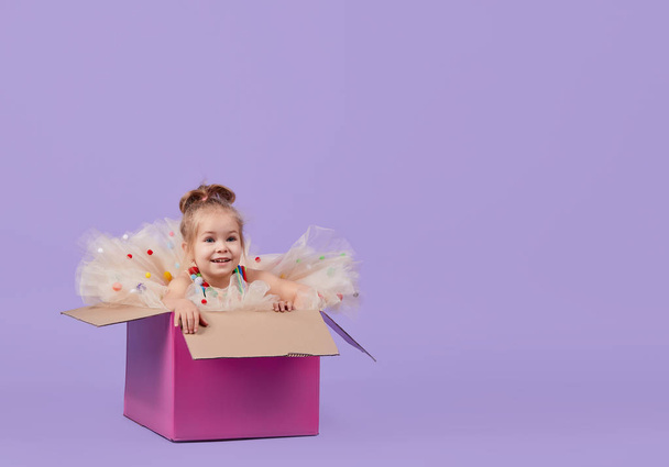 A cheerful little girl child in a magnificent holiday dress sits inside a large gift colored box, on a purple isolated background. Birthday party surprise. Space for text - Foto, Imagem