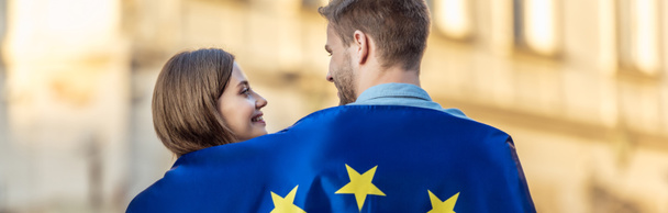 panoramic shot of young couple of tourists, wrapped in flag of european union, looking at each other on street - Foto, afbeelding