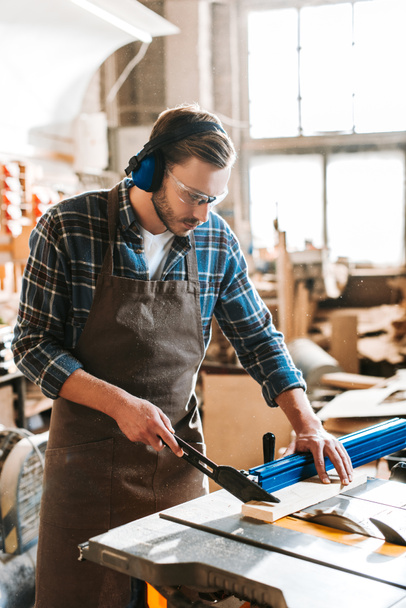 bearded woodworker in protective headphones and apron holding plank near circular saw - Photo, Image