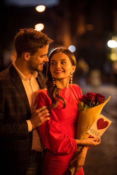 улыбающийся мужчина обнимает счастливую девушку, держа букет роз и открытку с надписью любви и символом сердца
 - Фото, изображение
