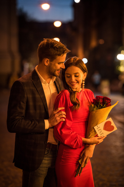 χαμογελαστός άνδρας αγκαλιάζει ευτυχισμένη φίλη κρατώντας μπουκέτο τριαντάφυλλα και κάρτα με την επιγραφή αγάπης και το σύμβολο της καρδιάς - Φωτογραφία, εικόνα