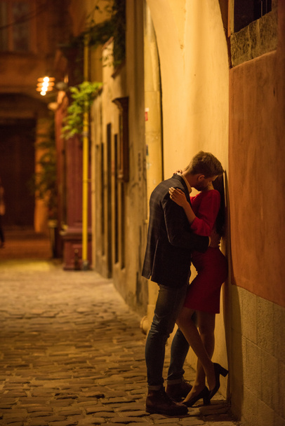 νεαρό ζευγάρι αγκαλιάζει και φιλιά, ενώ στέκεται κοντά στον τοίχο τη νύχτα - Φωτογραφία, εικόνα