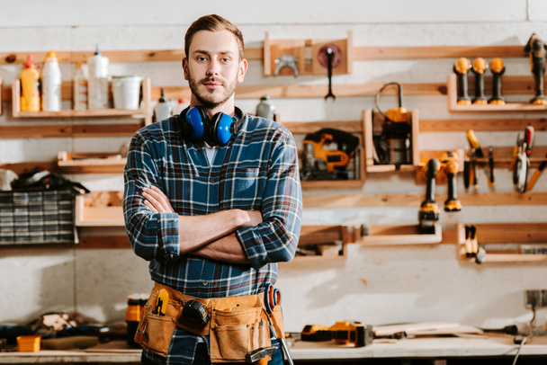 handsome carpenter in apron standing with crossed arms in workshop  - Fotoğraf, Görsel
