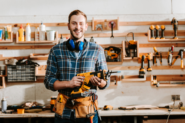happy bearded carpenter holding hammer drill in workshop  - Photo, Image