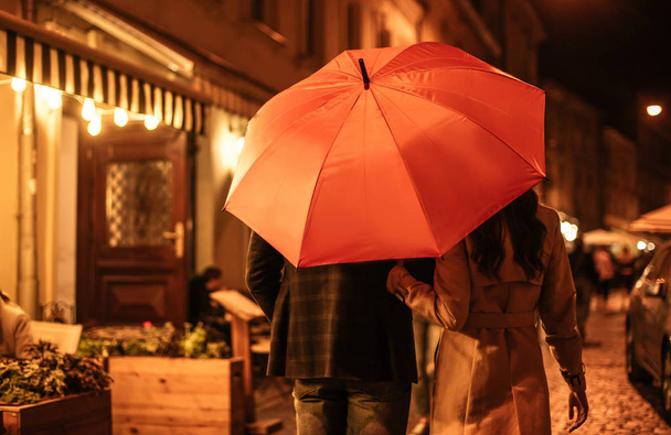 achteraanzicht van paar in herfst outfit wandelen onder paraplu langs avond straat - Foto, afbeelding