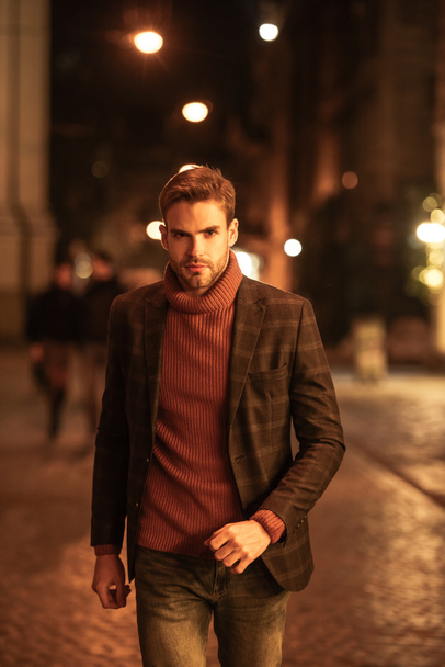 schöner, selbstbewusster Mann im Herbst-Outfit, der beim Spaziergang auf der Abendstraße in die Kamera schaut - Foto, Bild