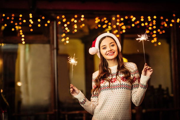 fröhliche junge Frau in warmem Pullover und Weihnachtsmütze mit Wunderkerzen in der Hand, während sie in die Kamera lächelt - Foto, Bild