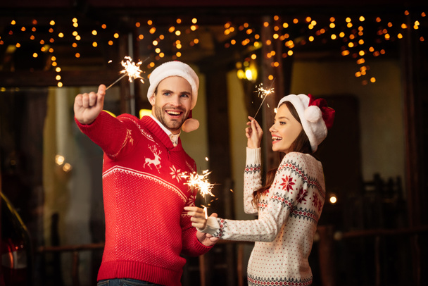happy young couple in santa hats and warm sweaters holding sparklers on christmas eve - Fotó, kép