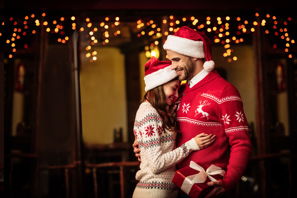 happy couple in warm sweaters and santa hats embracing on street - Foto, imagen