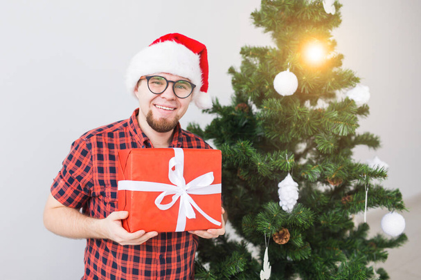 Christmas and holidays concept - Funny man in santa hat holding a gift at home in the living room - Fotografie, Obrázek