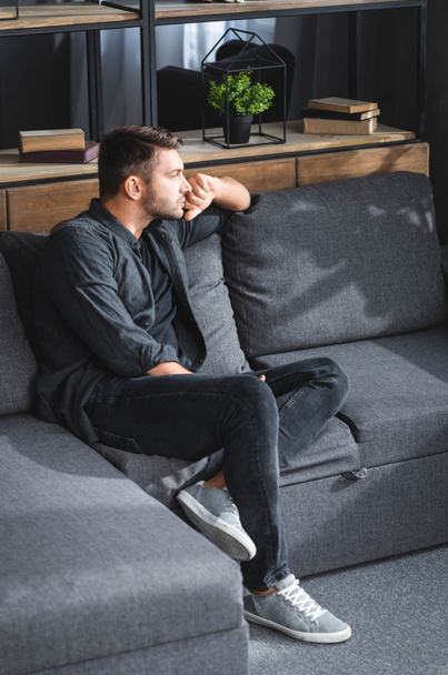 vysoký úhel pohledu pohledný a zamyšlený muž sedí na pohovce v bytě  - Fotografie, Obrázek