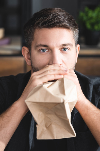 handsome man with panic attack breathing in paper bag in apartment  - Photo, Image