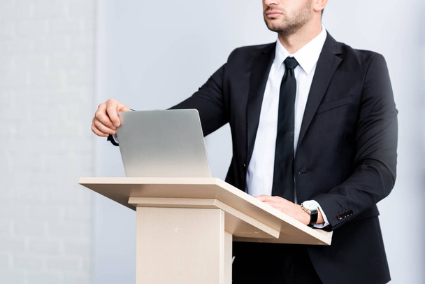 ostříhaný pohled na obchodníka v obleku stojící na pódiu tribune a otevírající notebook během konference izolované na bílém - Fotografie, Obrázek