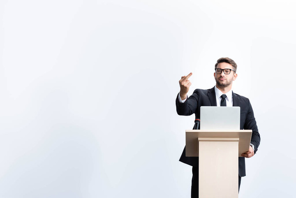businessman in suit standing at podium tribune and showing middle finger during conference isolated on white - Foto, afbeelding