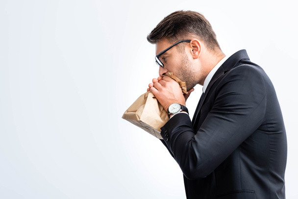 πλαϊνή άποψη του φοβισμένου επιχειρηματία με κοστούμι αναπνοή σε χάρτινη σακούλα κατά τη διάρκεια συνεδρίου απομονώνονται σε λευκό  - Φωτογραφία, εικόνα