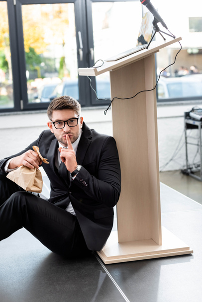 τρομαγμένος επιχειρηματίας με κοστούμι κρατώντας χαρτοσακούλα και δείχνοντας shh χειρονομία κατά τη διάρκεια του συνεδρίου - Φωτογραφία, εικόνα