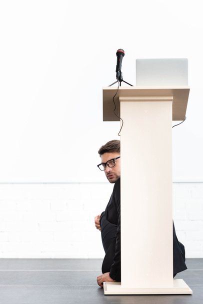 φοβισμένος επιχειρηματίας με γυαλιά κρύβεται πίσω από βάθρο tribune κατά τη διάρκεια συνεδρίου απομονώνονται σε λευκό  - Φωτογραφία, εικόνα