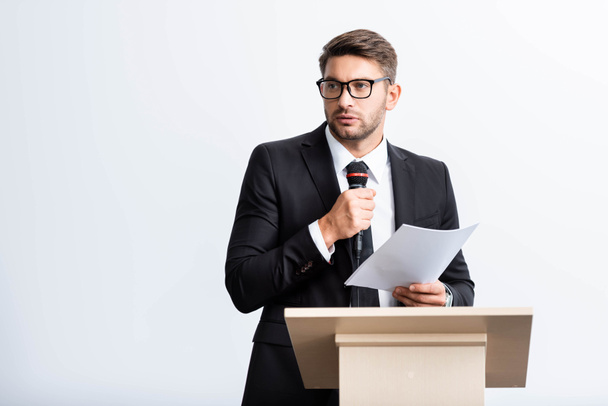 puvussa oleva liikemies seisoo puhujakorokkeella ja puhuu konferenssissa valkoisella eristyksissä
  - Valokuva, kuva