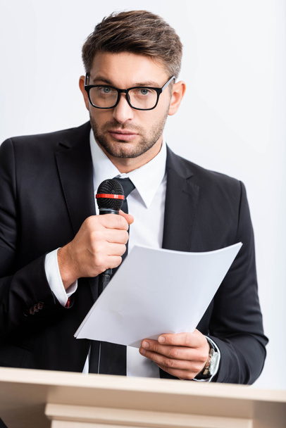επιχειρηματίας με κοστούμι κρατώντας χαρτί και μιλώντας κατά τη διάρκεια συνεδρίου απομονώνονται σε λευκό  - Φωτογραφία, εικόνα