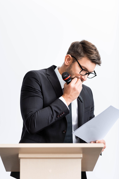испуганный бизнесмен в костюме стоит на трибуне и смотрит на бумагу во время конференции изолированы на белом
  - Фото, изображение