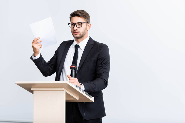 φοβισμένος επιχειρηματίας με κοστούμι στέκεται στο βάθρο tribune και κρατώντας μικρόφωνο κατά τη διάρκεια συνεδρίου απομονώνονται σε λευκό  - Φωτογραφία, εικόνα