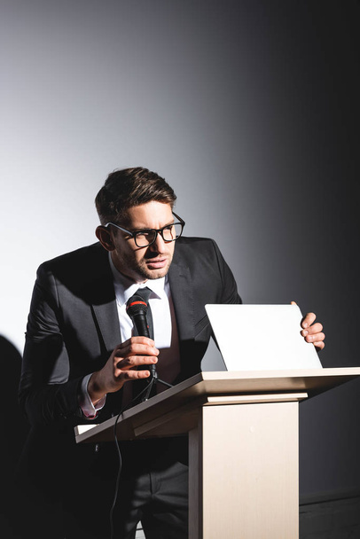 scared businessman in suit standing at podium tribune and holding laptop during conference on white background  - 写真・画像
