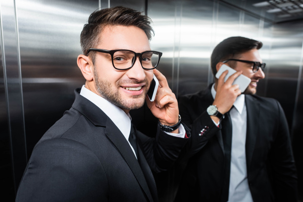 Lächelnder Geschäftsmann im Anzug spricht im Fahrstuhl auf Smartphone - Foto, Bild