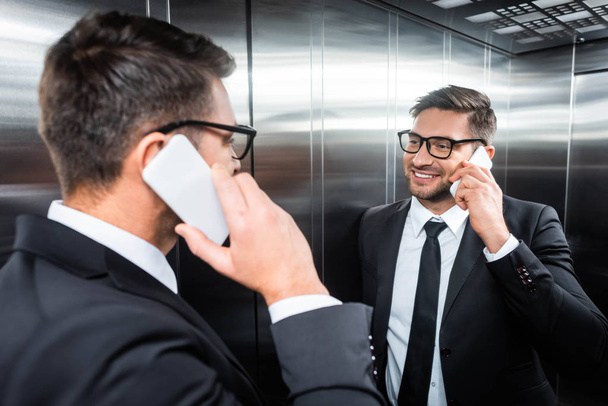 smiling businessman in suit talking on smartphone and looking at mirror in elevator - Fotó, kép
